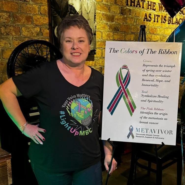 MaryAnn in a #LightUpMBC shirt beside a METAvivor 'Colors of the Ribbon' sign
